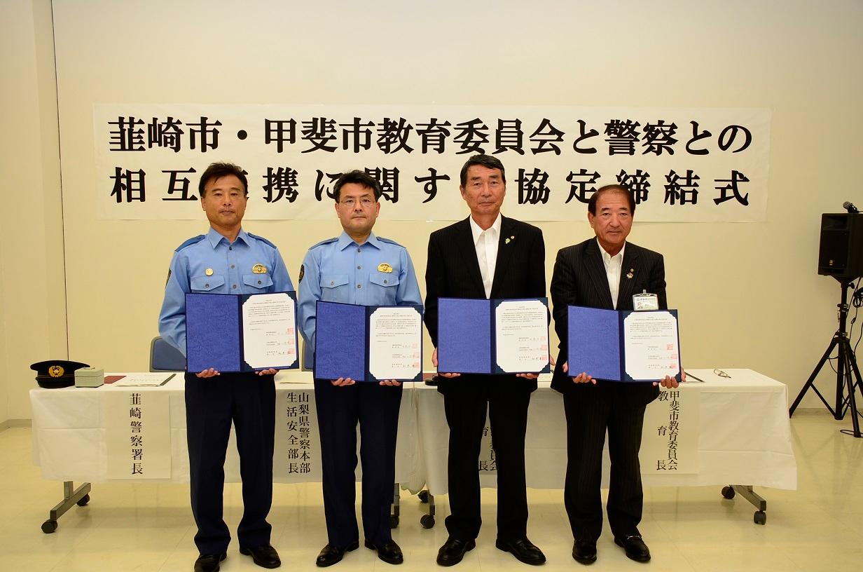 「韮崎市教育委員会と警察との相互連携に関する協定締結式」と書かれている横断幕の前で、協定書を手に持って、警察の関係者の男性2名と教育委員会の関係者の男性2名が並んで写っている写真