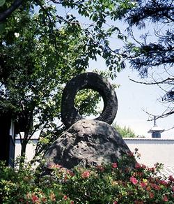 岩の上に中央部分がくり貫かれた円形の岩が乗せられており、岩の周りにはつつじの花が咲いている鏡石の写真