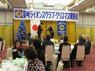 韮崎ライオンズクラブ・クリスマス家族会の横断幕の下の舞台上で、市長が黒いスーツの男性より寄付金を頂いている様子を円卓に座っている出席者の後方より撮影している写真