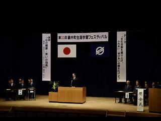 生涯学習フェスタの会場で、舞台上には「藤井町生涯学習フェスティバル」の横断幕と公園のテーマの垂れ幕が下がっており、壇上に男性が立って話をしている開会式の様子の写真
