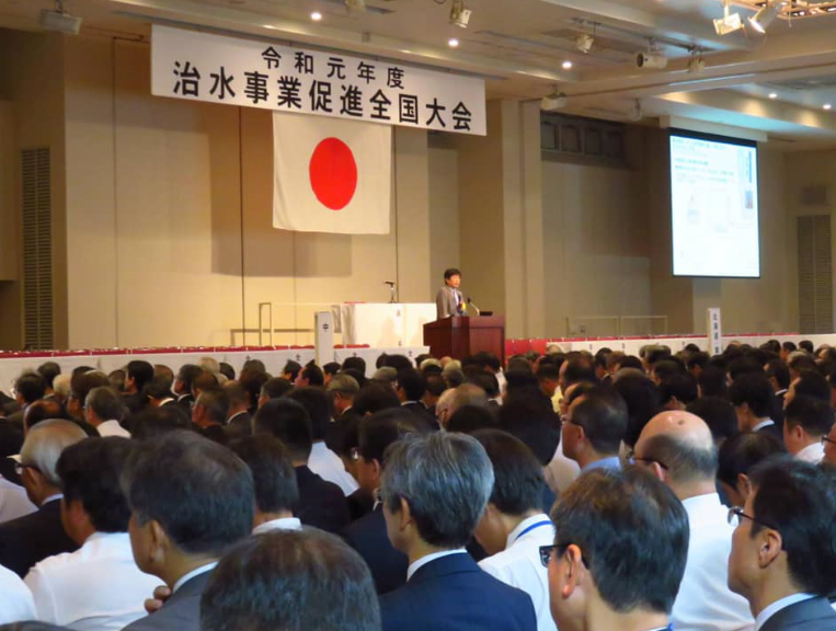 令和元年度治水事業促進全国大会で、演台に立つ男性と、前方を見ている関係者の写真