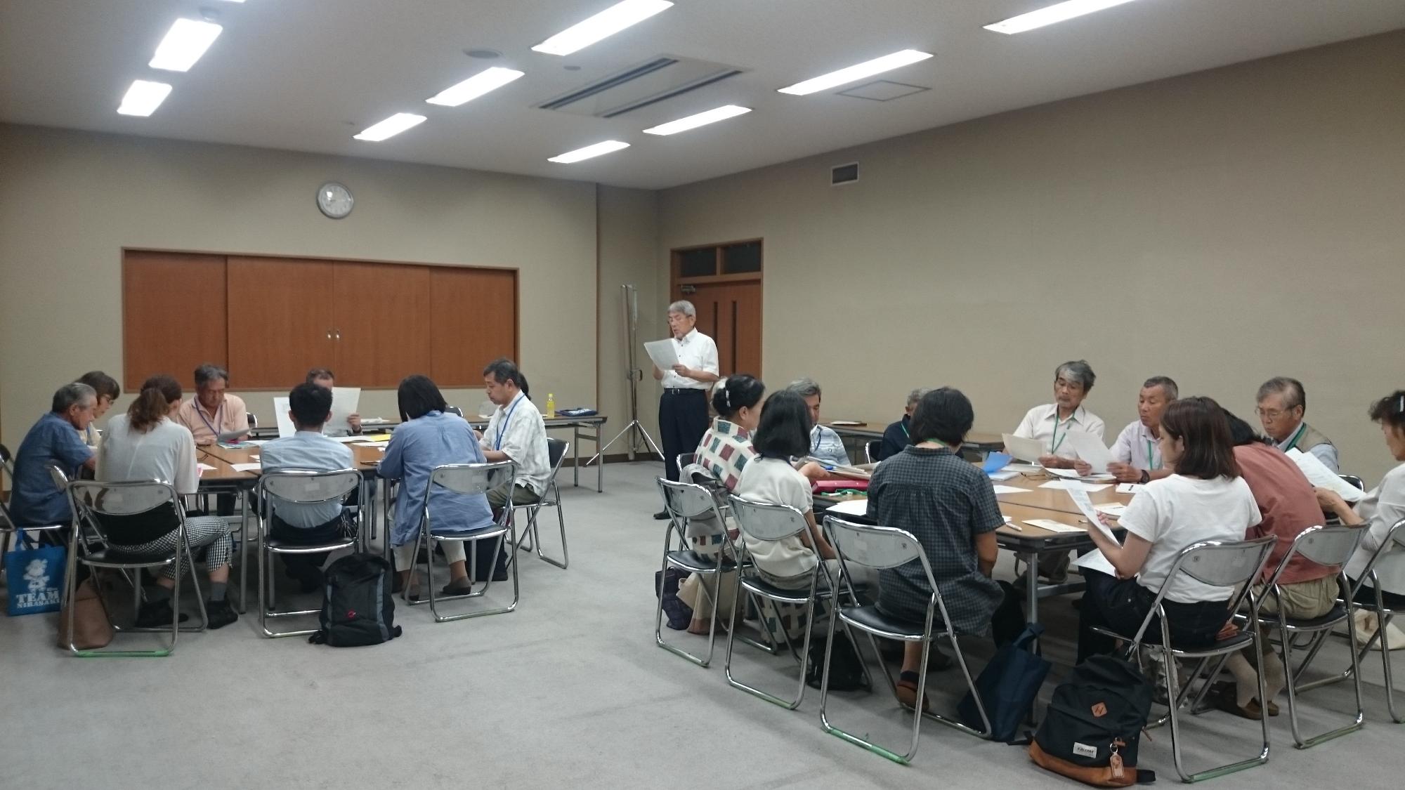 男性の方が室内の前方に立ち話をし座席についている人たちが手元の資料を見ている写真