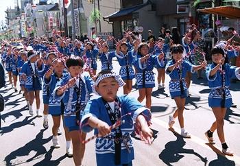 青い法被を着て、頭には豆絞りを巻いた子供たちが、通りで列を作って、手に紅白の綾棒を持って綾棒躍りを踊っている様子の写真