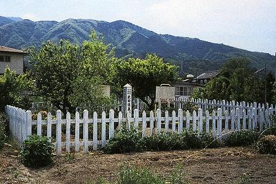 奥には山脈が見えており、畑の中に白い柵で囲まれている武田 信義館跡の遠景の写真