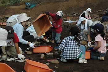 女夫石遺跡で親子連れ来た方々などがオレンジ色のテミを側に置いて、土を掘って発掘調査を体験している様子の写真