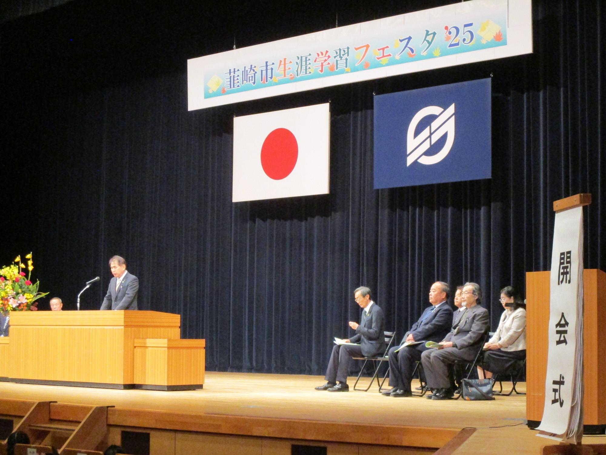 壇上に「生涯学習フェスタ&rsquo;」の横断幕が掲げられており、壇上に男性が立って話をしており、開会式が行われている様子の写真