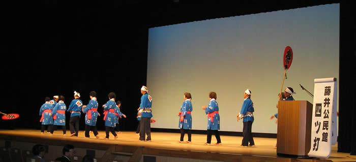 舞台の上で、「祭」と書いてある、大きなうちわを持った人や、青い法被を着た人々が、列を作って、四ツ打の踊りを踊って発表している様子の写真