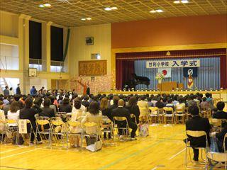 甘利小学校入学式の会場の舞台には「甘利小学校入学式」の横断幕と日の丸の国旗。校章の旗が掲げてあり、新一年生が会場の前方の椅子に座っており、その後ろの保護者席に保護者の方々が座っている入学式の様子の写真