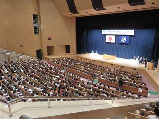 「武田の里ライフカレッジ」の開講式の様子を会場の上階より、満席の客席と舞台を写している写真