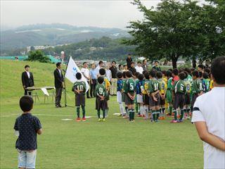 サッカー大会開催で、ユニホームを着た選手の皆さんがチーム毎に整列して並んでおり、市長がスタンドマイクの前に立って、選手の皆さんに話をしている様子の写真