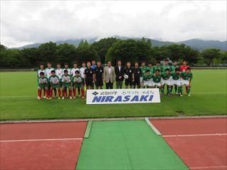 「武田の里 NIRASAKI」と書かれている横断幕の前で、サッカーフェスティバルに参加する選手の皆さんと関係者の方々がグラウンドで写っている集合写真