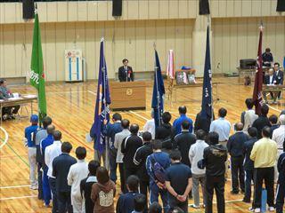 韮崎市体育祭り総合開会式で、チームの旗を持った選手を先頭に、チーム毎に整列しており、市長が壇上に立って話をしている様子の写真