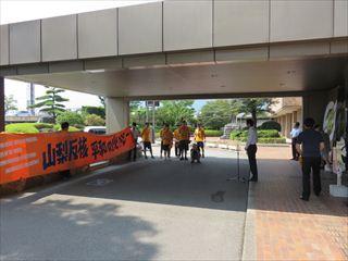 「山梨反核 平和の火リレー」と書かれているオレンジ色の横断幕が貼られており、市長がスタンドマイクの前に立って話をしている様子の写真