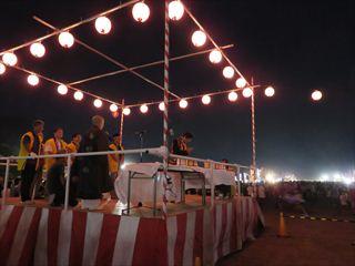 夏の武田の里まつりの会場で、舞台の上に提灯が四方に付けられて、明かりがともされており、祭りの進行が行われている様子の写真