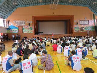 韮崎小学校の体育館で防災訓練が行われており、訓練スタッフと書かれているゼッケンを着ている関係者の方や、市民の皆様が大勢集まっており、床に座って説明を聞いている様子の写真