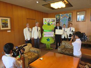 カメラマンの前で、ニーラーと横で賞状をもつ市長、関係者がならんでいる写真