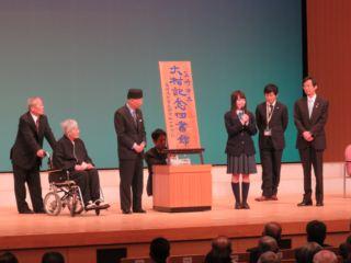 「韮崎市立大村記念図書館」の刻字看板の横でマイクをもって話す女子学生と、その横に並ぶ関係者の写真