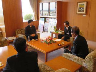 応接室に加賀美 孝久さんが市長の左側に座っており、笑顔で談笑している様子の写真