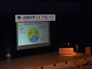 武田の里ライフカレッジでスクリーンを使って地域の歴史や市の財政について講演をしている市長の写真