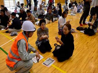 防災訓練で、韮崎西中学校の体育館に集まる住民と、ヘルメットをかぶり作業着の市長が話をしている写真