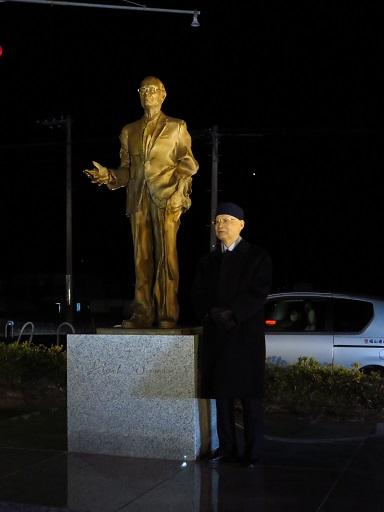 ライトアップされる大村智博士の銅像の横に立つ男性の写真