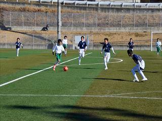 白いユニホームを着た選手がボールを蹴って走っており、相手チームの青いユニホームを着た選手たちがボールを狙って追いかけている様子のサッカーの試合の写真