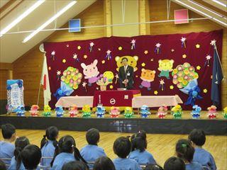 可愛い動物たちやお花で壁面が飾られている舞台の上で、市長が園児の皆さんに話をしており、園児の皆さんが真剣に話を聞いている様子を園児の皆さんの後方より写している写真