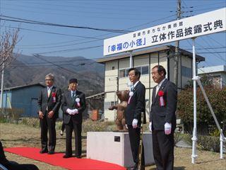 赤い絨毯の中央にクマのような小径立体作品が置かれており、その横に市長、大村 智博士,関係者が一緒に並んでおり、小径立体作品の除幕式が行われている様子の写真