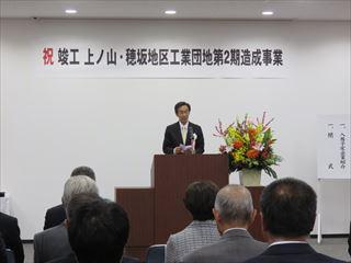 「祝竣工 上ノ山・穂坂地区工業団地 第2期造成事業」と書かれている横断幕の前で、市長が壇上に立って関係者の皆さんに話をしている様子の写真