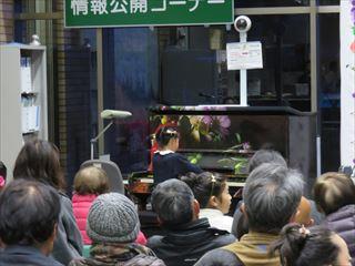 市役所のロビーに置かれているピアノに女の子が座って演奏しているのを、沢山のお客さんが見ている様子を後方から写している写真