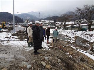 雪が薄っすらと降り積もっている土石流の災害現場に市長と関係者の方々が、ヘルメットと防寒着を着て視察をしている様子の写真