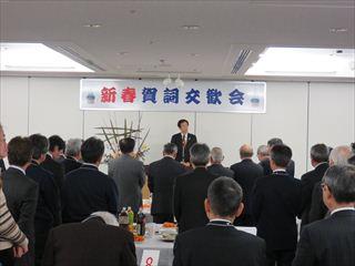 「新春賀詞交歓会」と書かれている横断幕の前で、市長が市内企業代表者の参加者の皆様の前で新年の挨拶をしている様子の写真