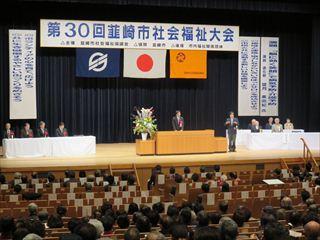 「第30回 韮崎市社会福祉大会」の横断幕の前で、市長が壇上で話をしており、客席には沢山のお客さんが席に着いている社会福祉大会の様子の写真