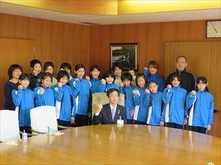 韮崎バレーボールスポーツ少年団の選手の皆さんが、青いジャンパーを着て、椅子に座っている市長の後ろに並んで、ガッツポーズをして写っている集合写真