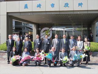 韮崎市役所の玄関の前で、退職者の職員の方々が花束を持っており、市長と関係者が一緒に並んで写っている集合写真