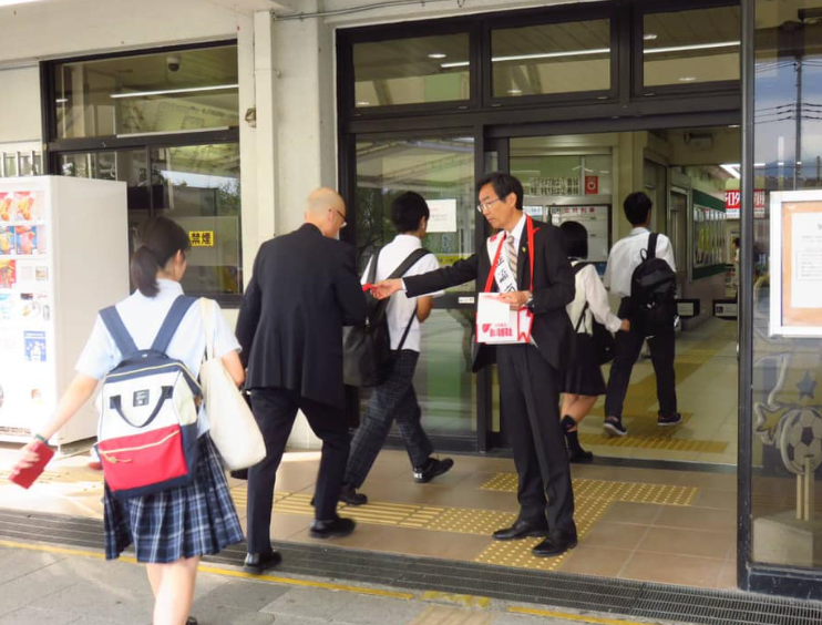 募金箱を首から下げた市長が、韮崎駅前で駅を利用している人々に赤い羽根を渡している写真