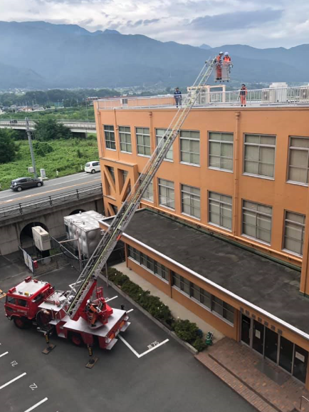 市役所の屋上に伸びる消防車のはしご車に乗る消防団員2名と、屋上にいる2名の関係者の写真