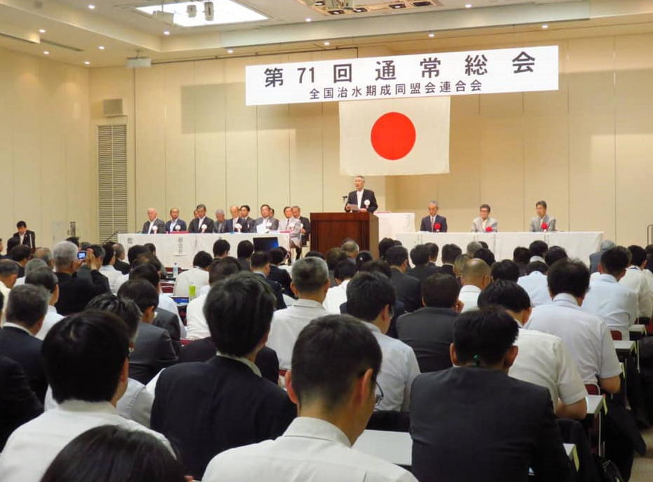 「第71回 通常総会」と書かれている横断幕の下に日の丸が掲げられており、壇上では男性が話をしており、舞台上の横に設けられた席に、胸に赤い花をつけた関係者の皆さんが座っており、たくさんの出席者が席に着いて話を聞いている総会の様子の写真