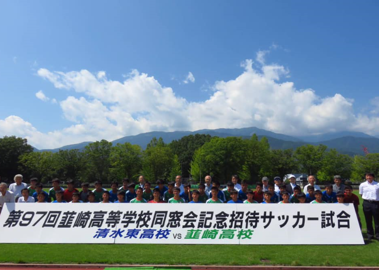 第97回韮崎高校同窓会記念招待サッカー試合と書かれたプレートの後ろに並ぶ、ユニフォームを着たサッカー部員と関係者の集合写真