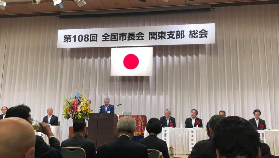 「第108回 全国市長会関東支部総会」と書かれている横断幕の下に日の丸が掲げらえており、壇上では男性が話をしており、壇上の横には席が設けられて胸に赤い花をつけた関係者の皆さんが座っており、たくさんの出席者が椅子に座って話を聞いている総会の様子の写真