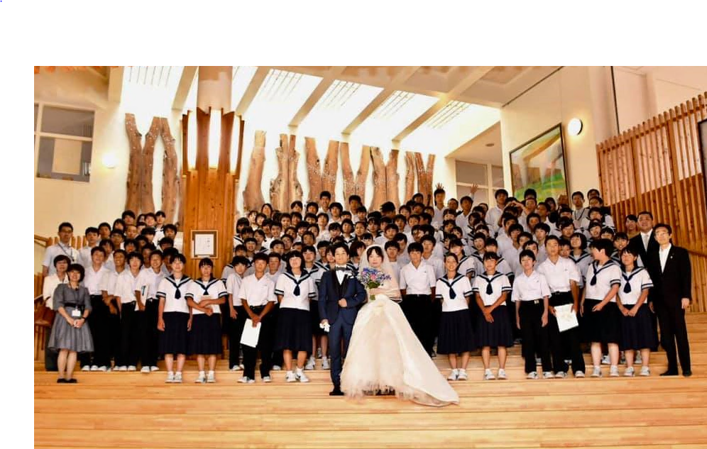 階段に並んで、白いウエディングドレスとタキシード姿の新郎新婦役の二人を中心に韮崎西中学校の生徒と一緒に写っている集合写真