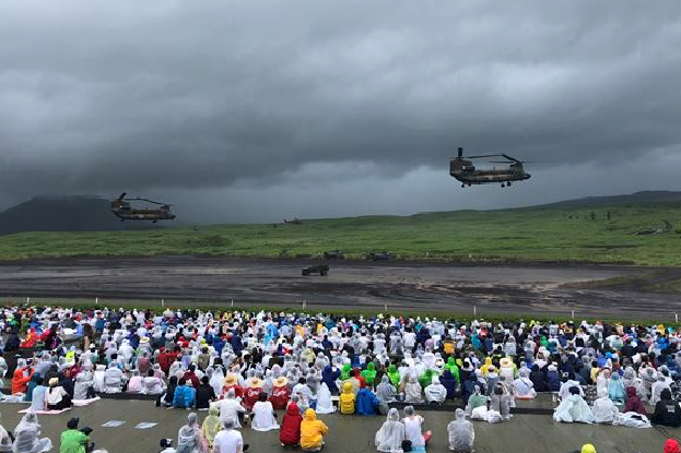 前方と後方に2つのプロペラが付いている2機のヘリコプターが飛んでいる様子を沢山の人が地面に座り見ている写真