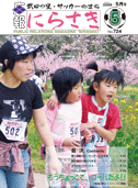 菜の花や桜の花が咲いている道をゼッケンを付け肩を組んで歩いている女の子二人と女性一人が写っている広報にらさき5月号表紙