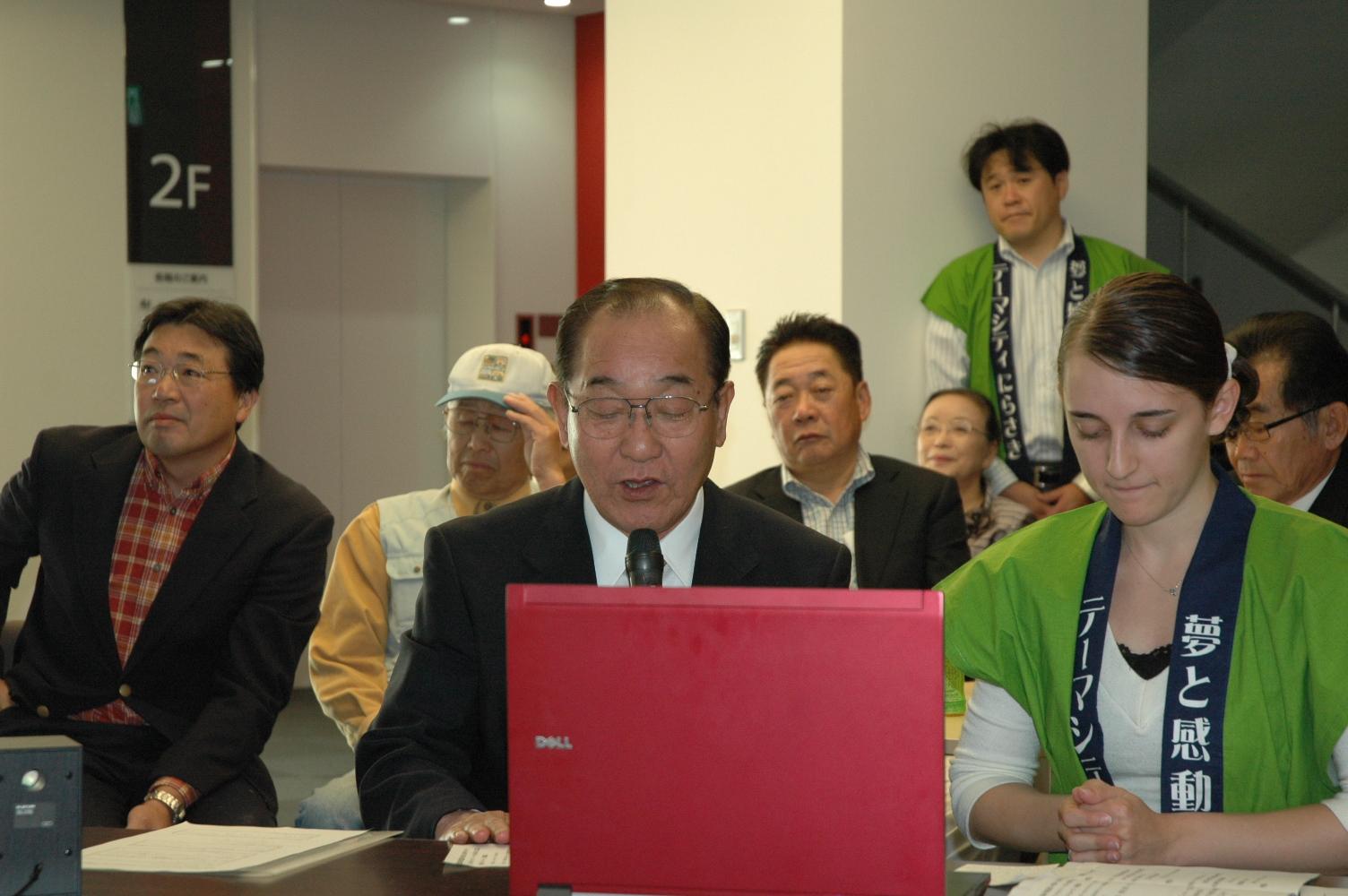 韮崎市長と緑色のはっぴを着た外国人女性がノートパソコンの前に座り、その後ろに座っている市民の人達の写真
