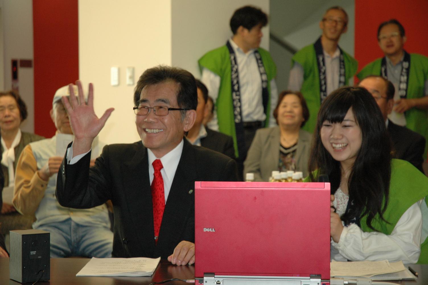 ノートパソコンの前に座っている緑色のはっぴを着た笑顔の女性と山寺 健二さんが笑顔で手を振っている写真