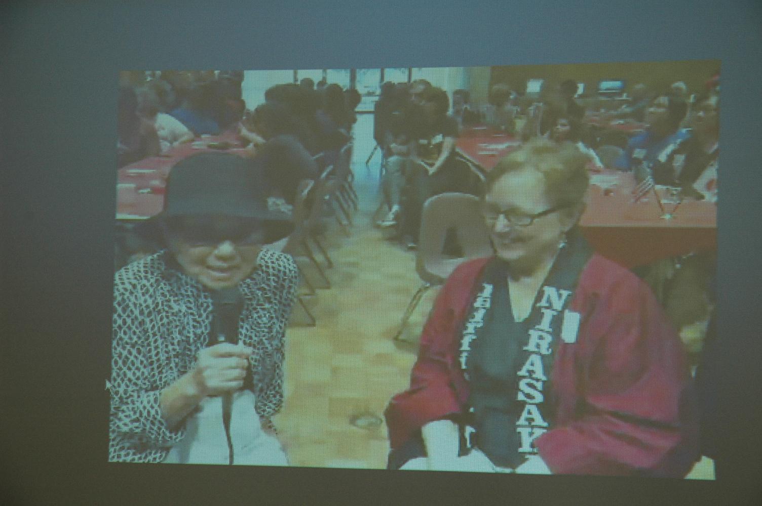 スクリーンに映し出された黒い帽子を被り眼鏡をかけ、手にマイクを持っているキエコ ウィタカーさんとアーレッタ コートライトさんの写真