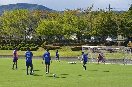 緑の芝生が広がっているグラウンドのゴール前でサッカーの練習をしている選手達の写真