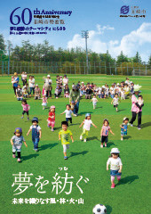 青空が広がる中、緑の芝生がはられたグラウンドで保護者の方と幼児が地面に座っていたりサッカーボールを追いかけてる子達の写真が載っている市制施行60周年記念市勢要覧の表紙